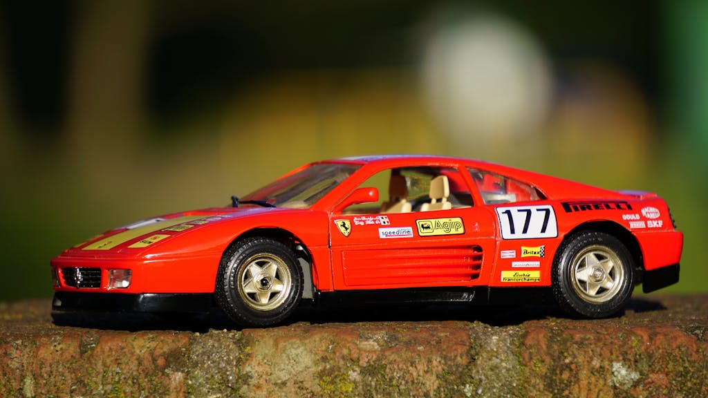 Close-up of a detailed red model sports car on a textured surface outdoors with a blurred background.